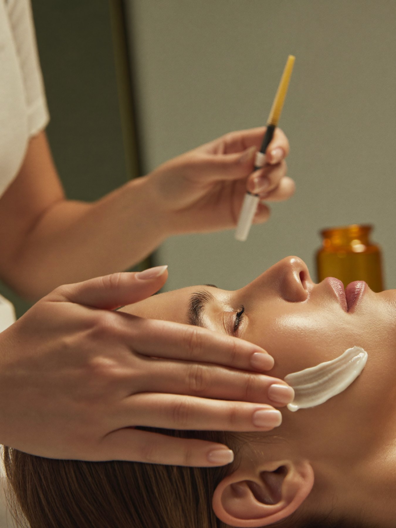Mãos da profissional aplicando cuidado facial com gesto preciso, frasco âmbar ao fundo