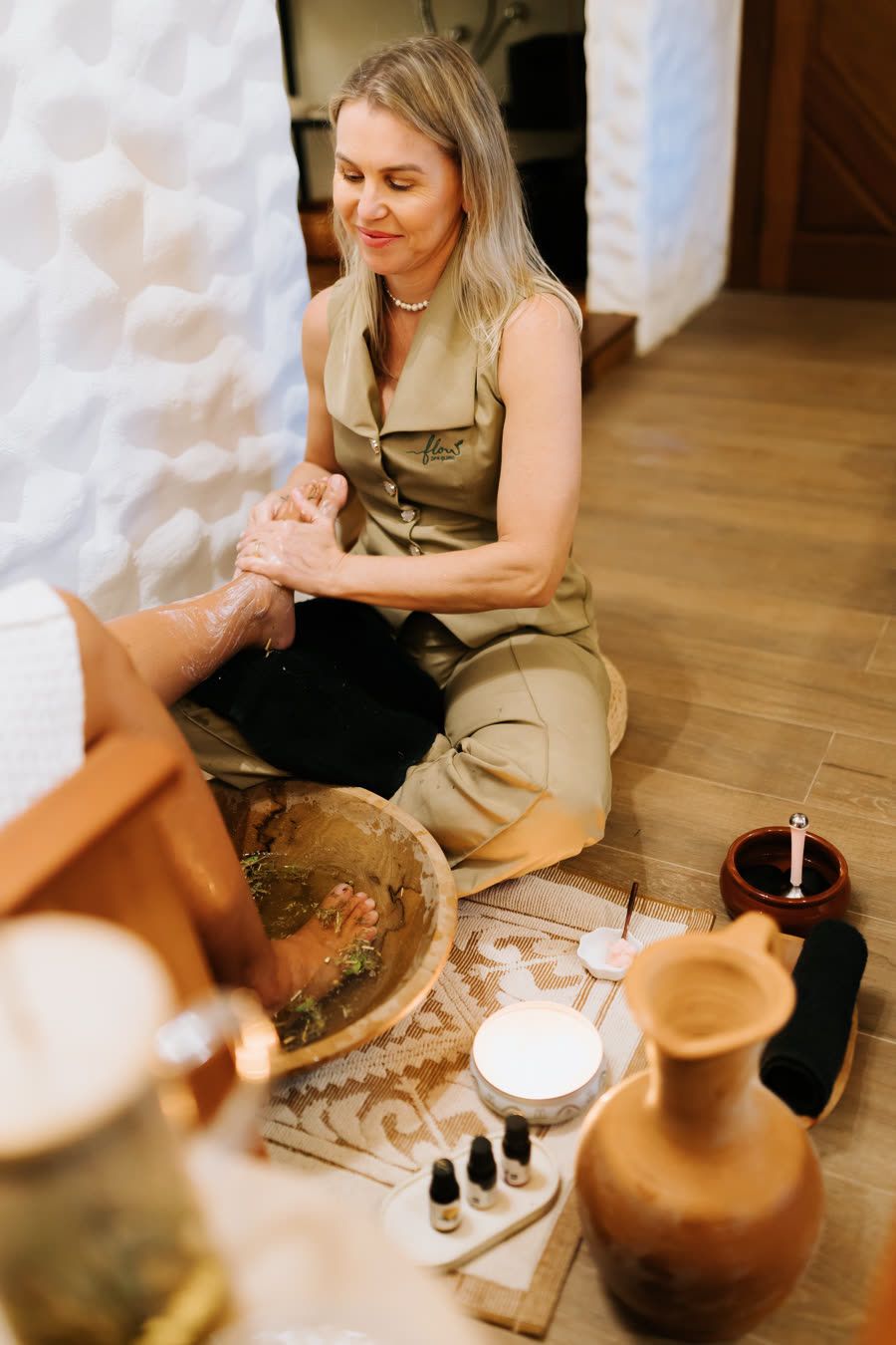 Terapeuta do Flow, sorrindo, lavando os pés da cliente no escalda-pés com ervas